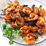 Spicy grilled shrimp skewers piled on a white platter with fresh parsley.