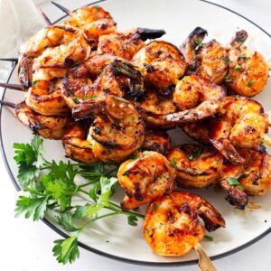 Spicy grilled shrimp skewers piled on a white platter with fresh parsley.