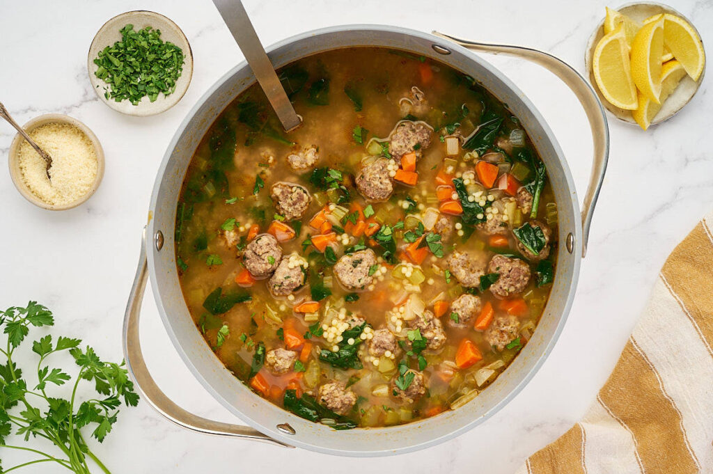 Pot of Italian wedding soup surrounded by bowls, lemon wedges, herbs, and serving spoons.