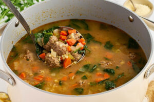 Large pot of Italian wedding soup with a ladle, meatballs, vegetables, greens, and small pasta.
