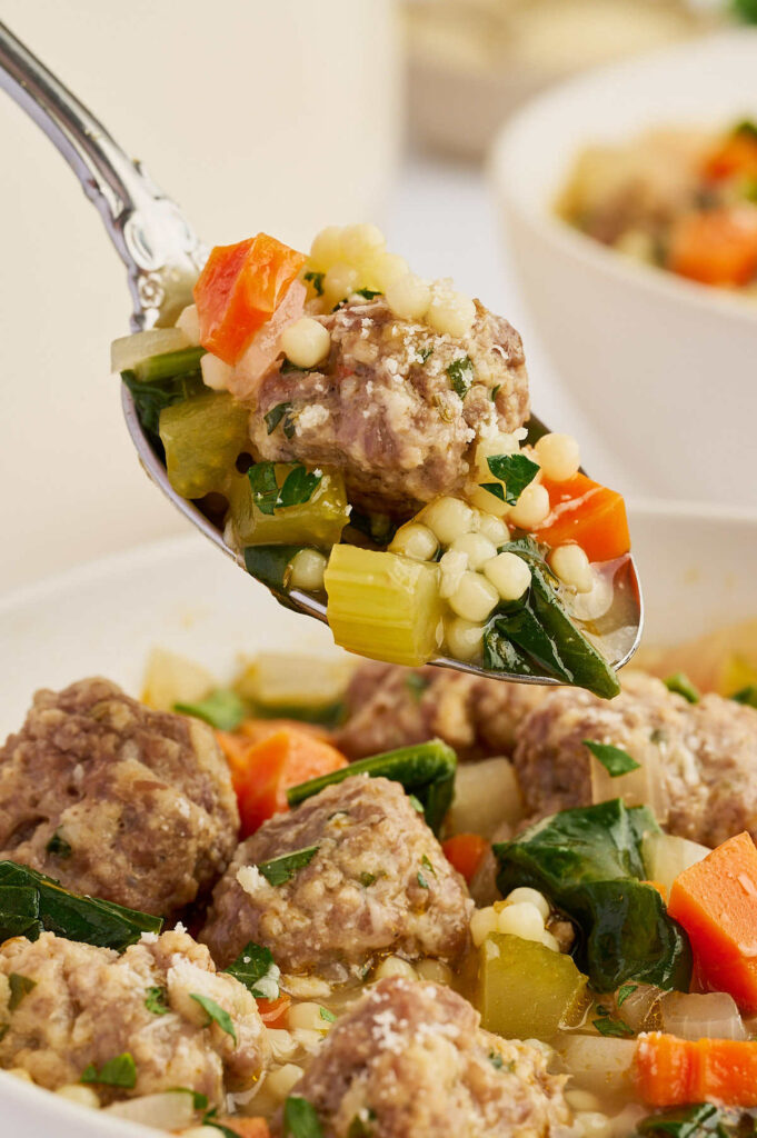 Close-up of Italian wedding soup with a spoon lifting a meatball, vegetables, and tiny pasta from the broth.
