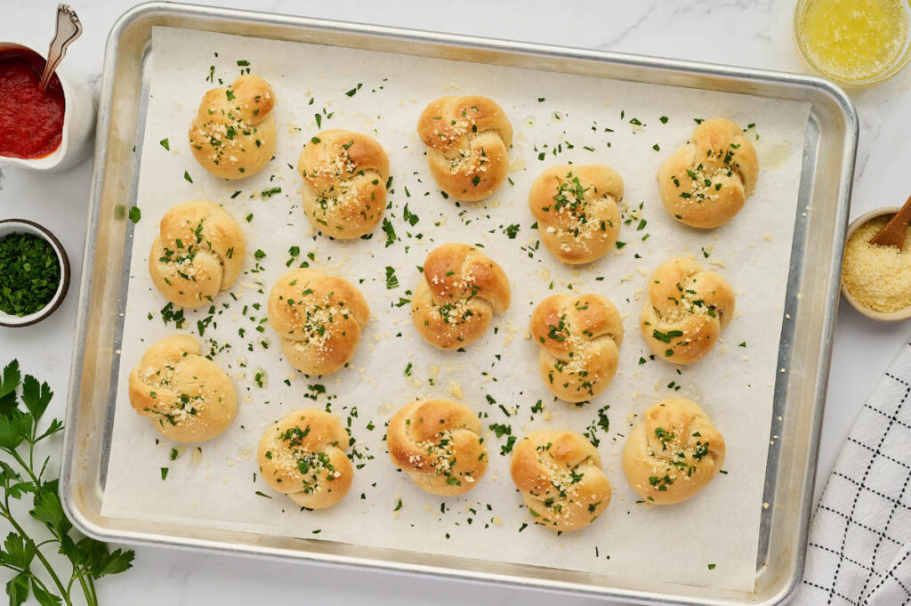 Baked homemade garlic knots on a sheet pan with marinara sauce and garlic butter.