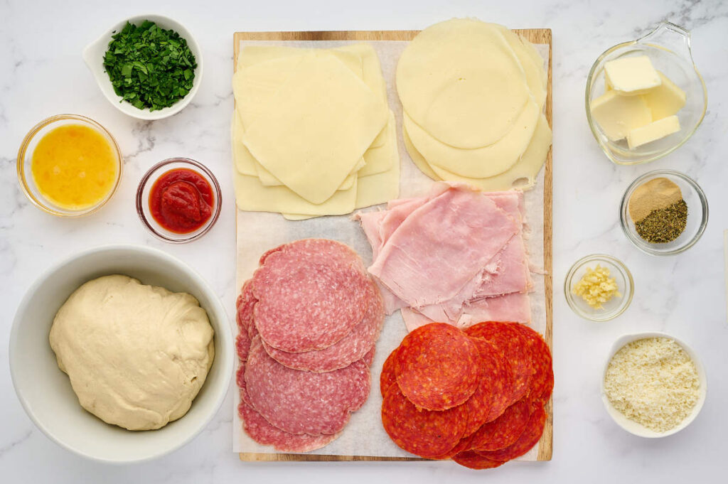 Ingredients for a meat stromboli arranged with pizza dough, cheeses, deli meats, and toppings.