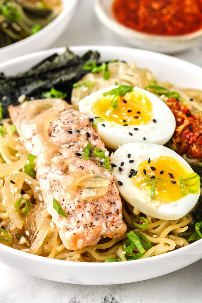 Close view of salmon ramen with soft-boiled eggs, noodles, sliced onions, nori, and chili crisp.