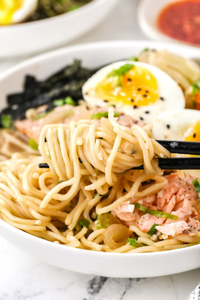 Chopsticks lifting noodles from a bowl of salmon miso ramen with soft-boiled egg and salmon in the background.