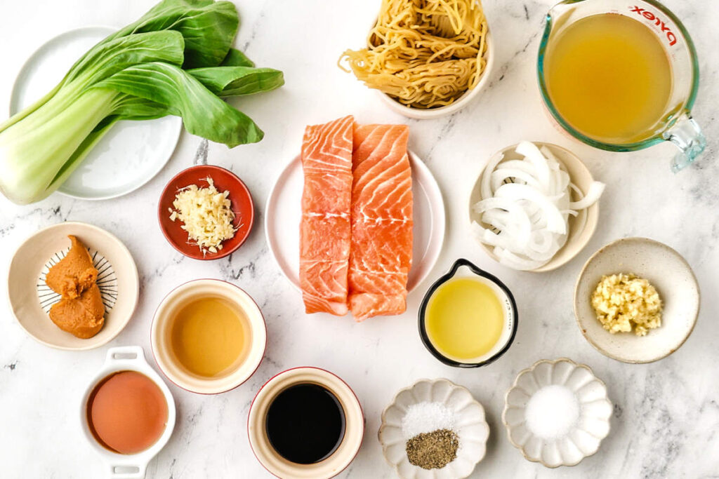 Ingredients for miso salmon ramen arranged with salmon fillets, bok choy, ramen noodles, broth, miso, soy sauce, ginger, and garlic.