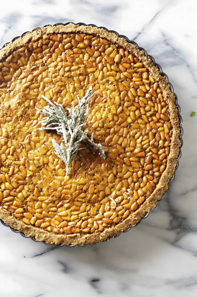 Pine nut tart with golden crust and sugared rosemary on marble surface.