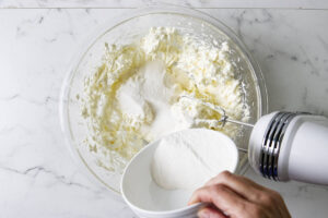 Sugar and flour mixture being added to softened cream cheese for pistachio Basque cheesecake.