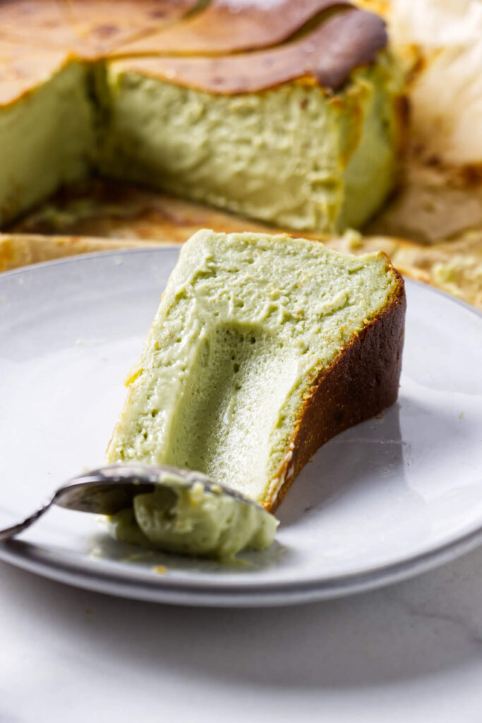 Pistachio Burnt Basque cheesecake slice on a plate with the whole cheesecake in the background.
