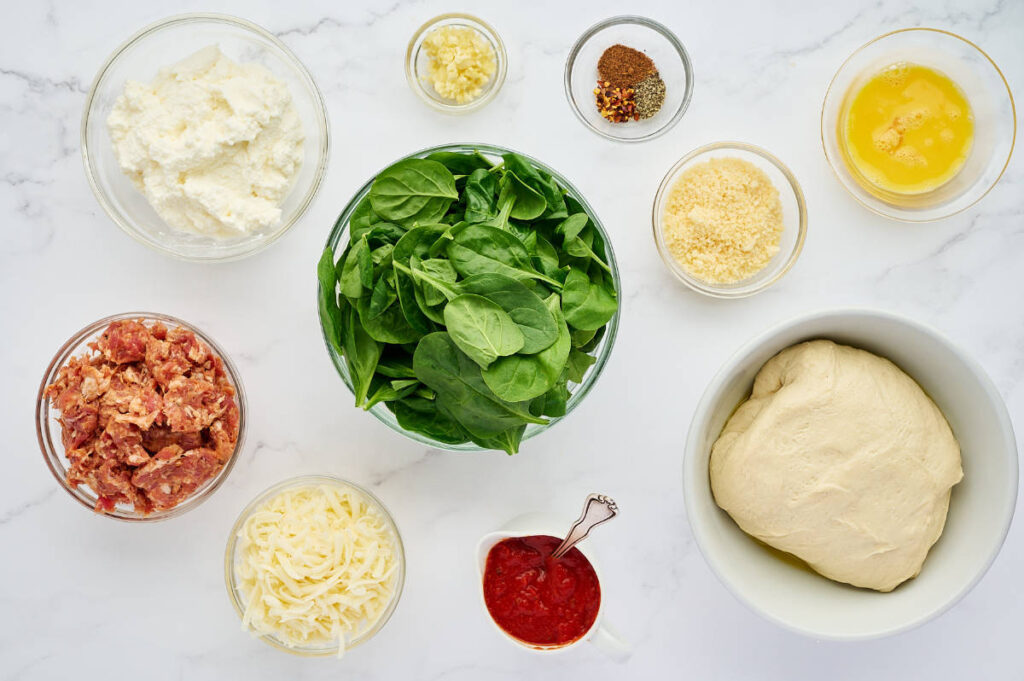 Ingredients for sausage ricotta calzones arranged with pizza dough, cheeses, sausage, spinach, and seasonings.