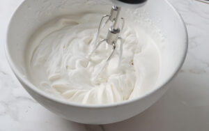 Electric hand mixer whipping cream in a white bowl, soft peaks forming.