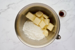 Metal mixing bowl with flour, cubed butter, and vanilla nearby.