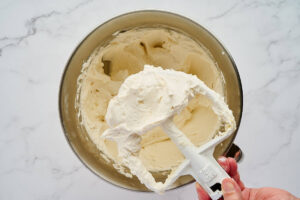 Hand holding a mixer attachment covered in fluffy white frosting.