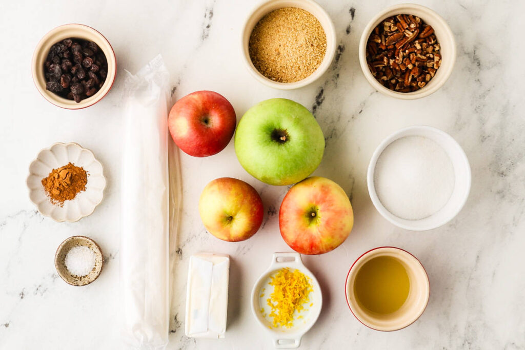 Apples, pastry dough, and small bowls of baking ingredients on a surface.