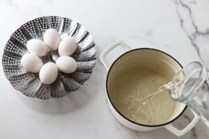Five raw eggs in a metal steamer basket near a pot.