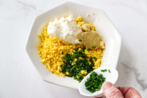 Hand adding chopped chives to mashed egg yolks in a bowl.