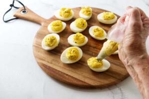 Hand piping yolk filling into halved hard-boiled eggs on a board.