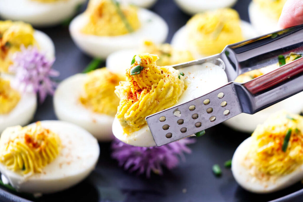 Metal tongs holding a deviled egg with paprika and chives.