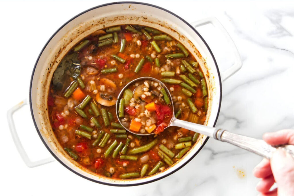 Metal ladle lifting vegetable barley soup from a white pot.