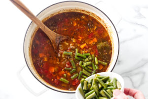Chopped green beans being added to a pot of vegetable soup.
