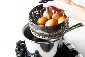 Hand lowering a steamer basket of brown eggs into a pot.