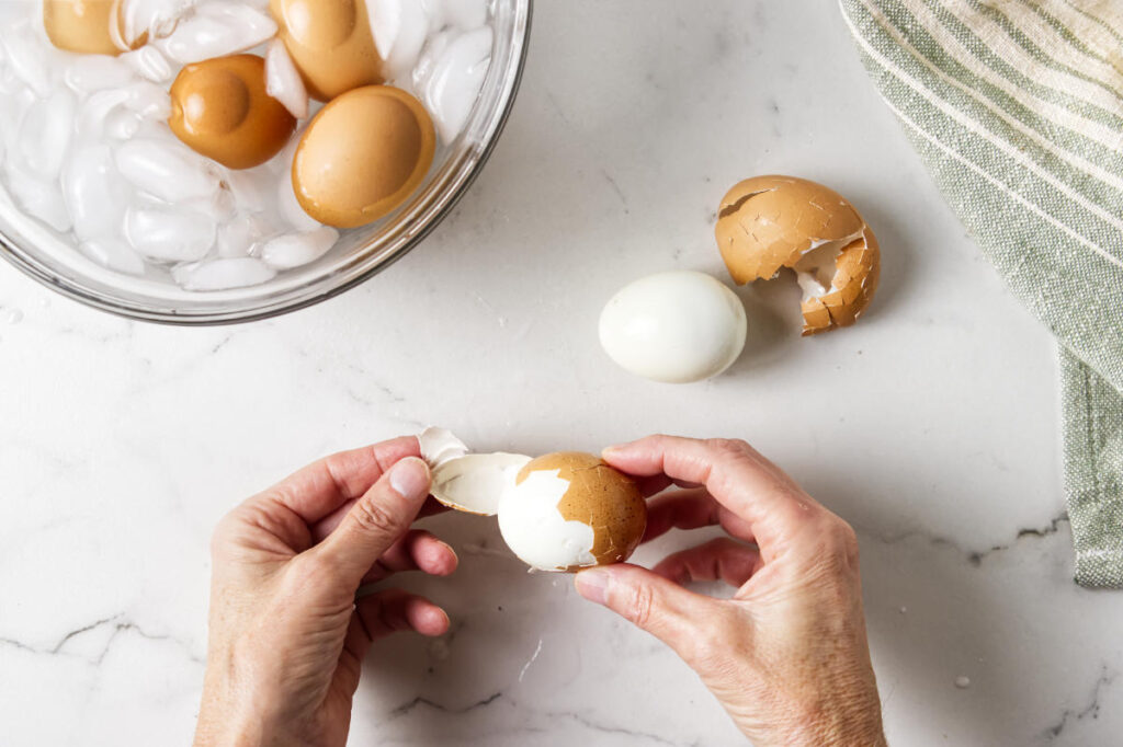 Hands peeling a hard-boiled egg next to a bowl of ice.